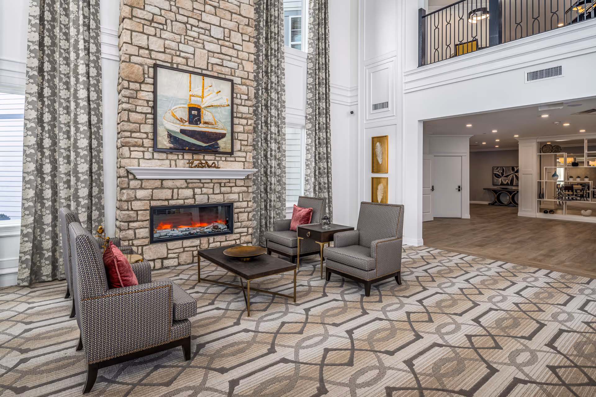 A spacious and elegant living room area with a tall stone fireplace featuring an electric fire and a painting of a boat above it. The room has large windows with patterned curtains, four upholstered armchairs with red accent pillows arranged around a dark coffee table, and a patterned carpet. The background shows an open hallway with white walls and wood flooring.