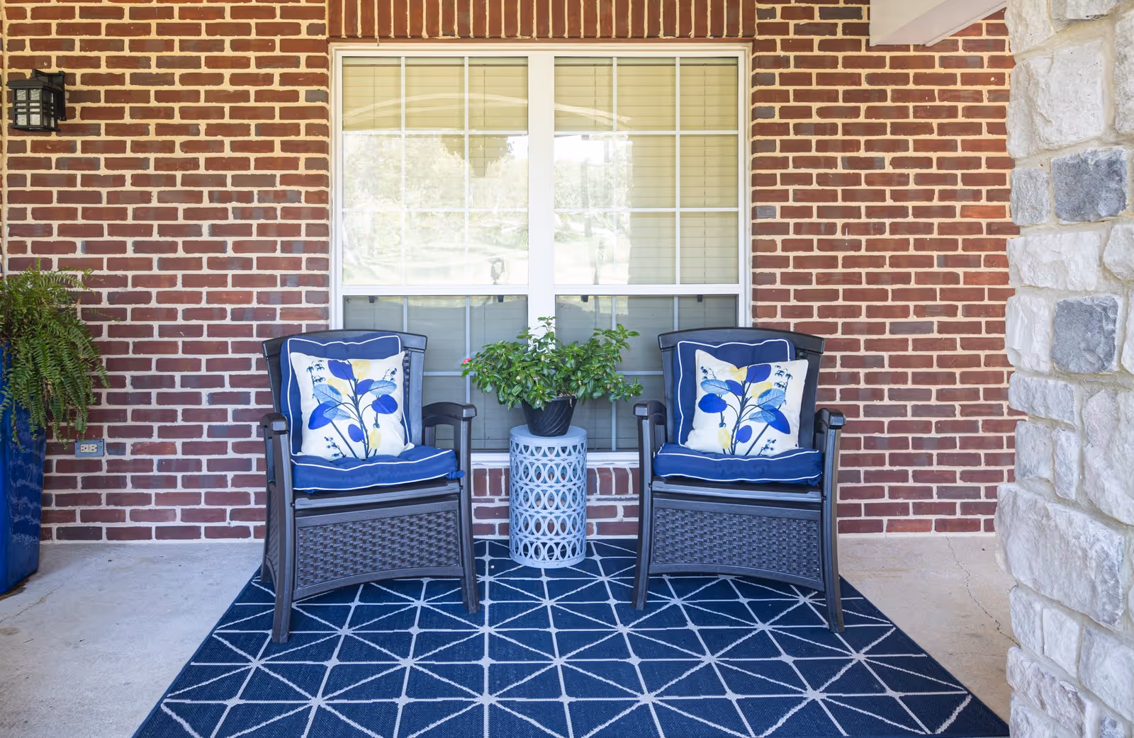 Two black wicker chairs with blue cushions and floral pillows sit on a blue geometric patterned outdoor rug in front of a brick wall with a large window. A small white decorative table with a green potted plant is placed between the chairs. A blue planter with a fern is visible on the left side, and a stone pillar is on the right.