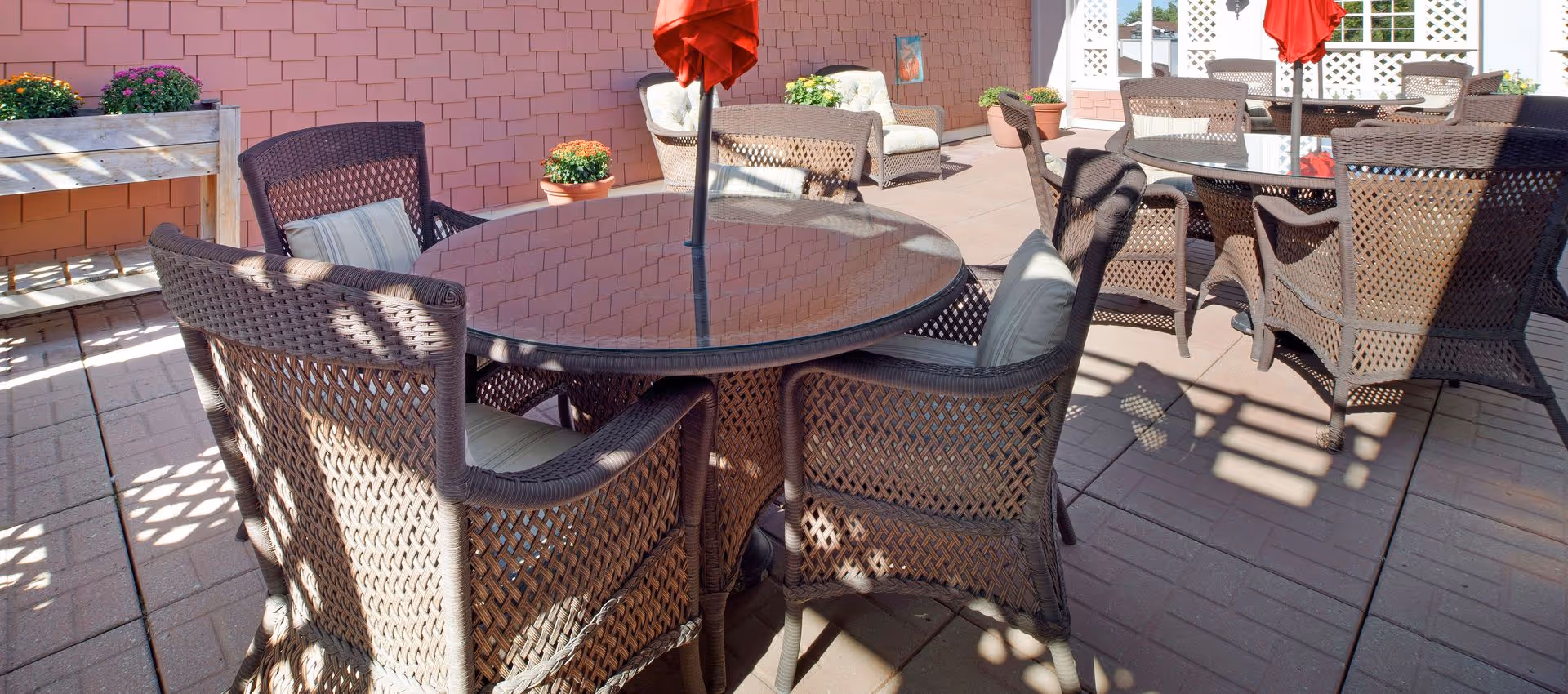 Outdoor patio with round glass-top tables, wicker chairs, red umbrellas, and potted plants on a brick paver surface.