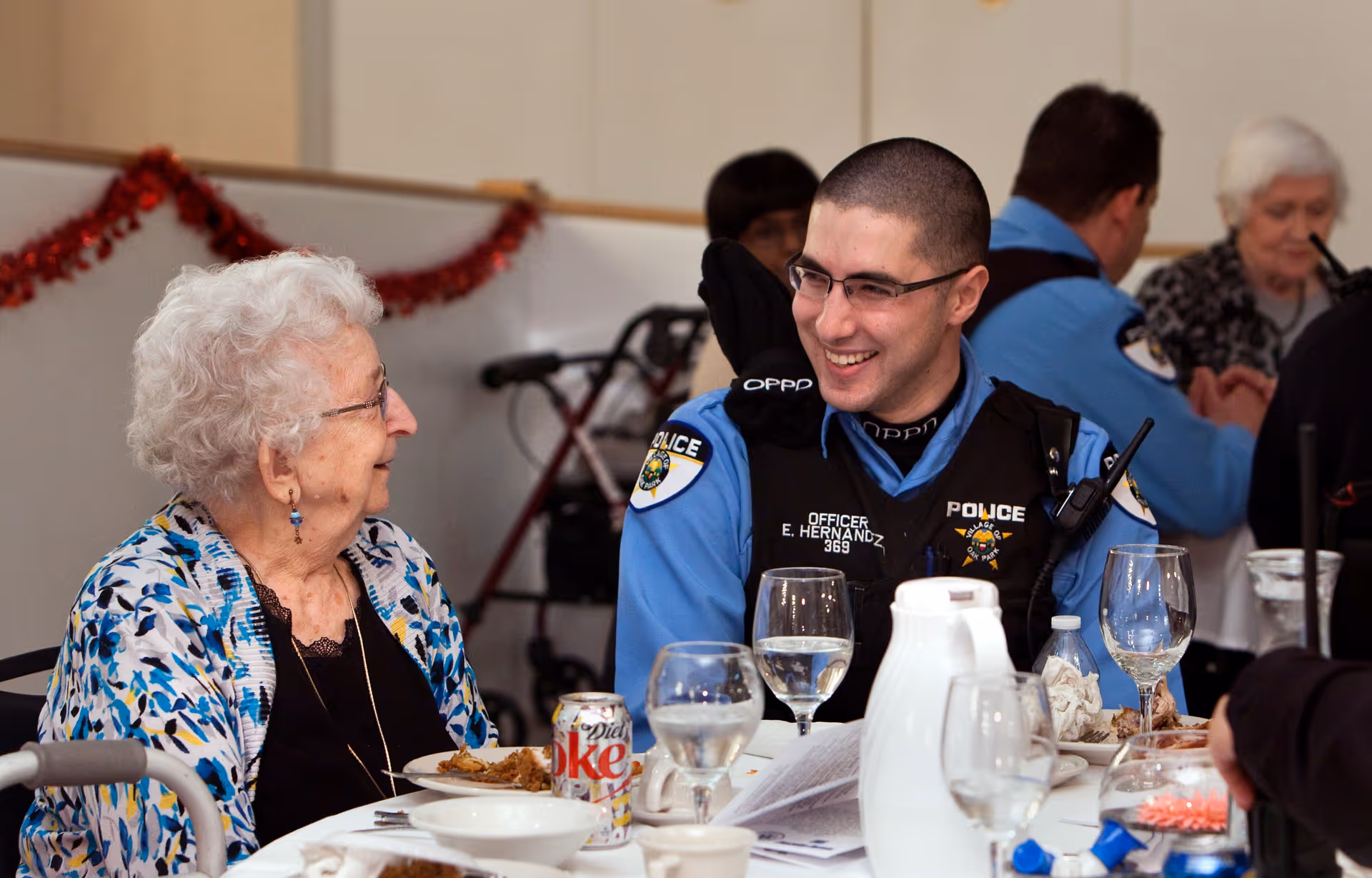 An elderly woman and a police officer in uniform are sitting at a dining table, smiling and engaging in conversation. The table has plates, glasses, a Diet Coke can, and a white pitcher. In the background, other people are seated and a walker is visible.