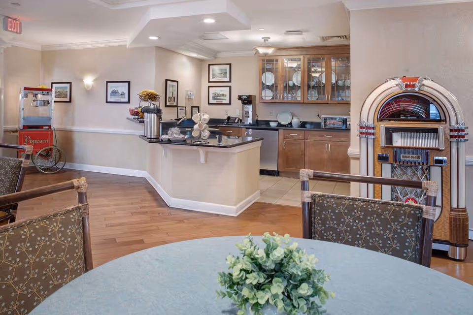 Community dining area with a round table and plant in the foreground, a serving counter and kitchenette, popcorn machine, and jukebox.
