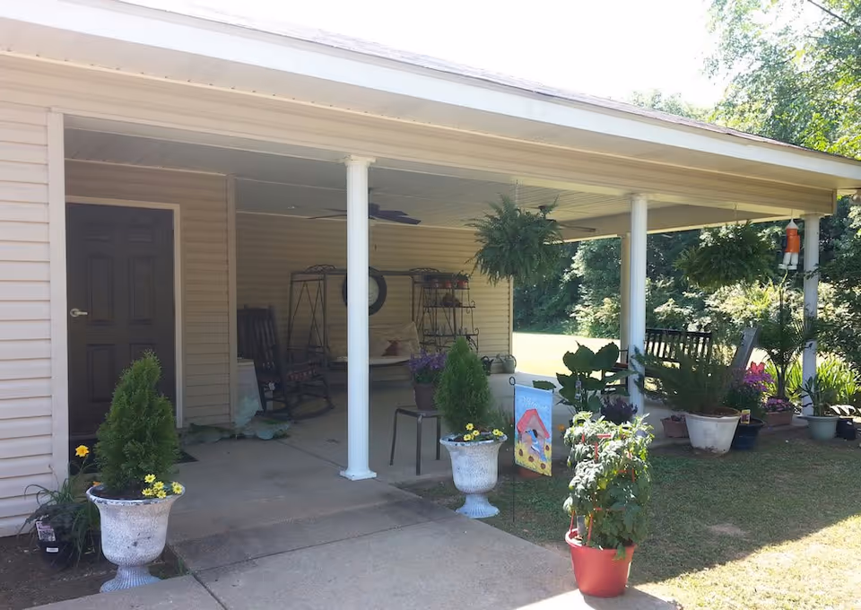 Covered outdoor patio area with white columns, several potted plants and flowers, rocking chairs, a swing bench, and ceiling fans. The patio is attached to a beige building with a dark door, and there is a grassy area and trees in the background.
