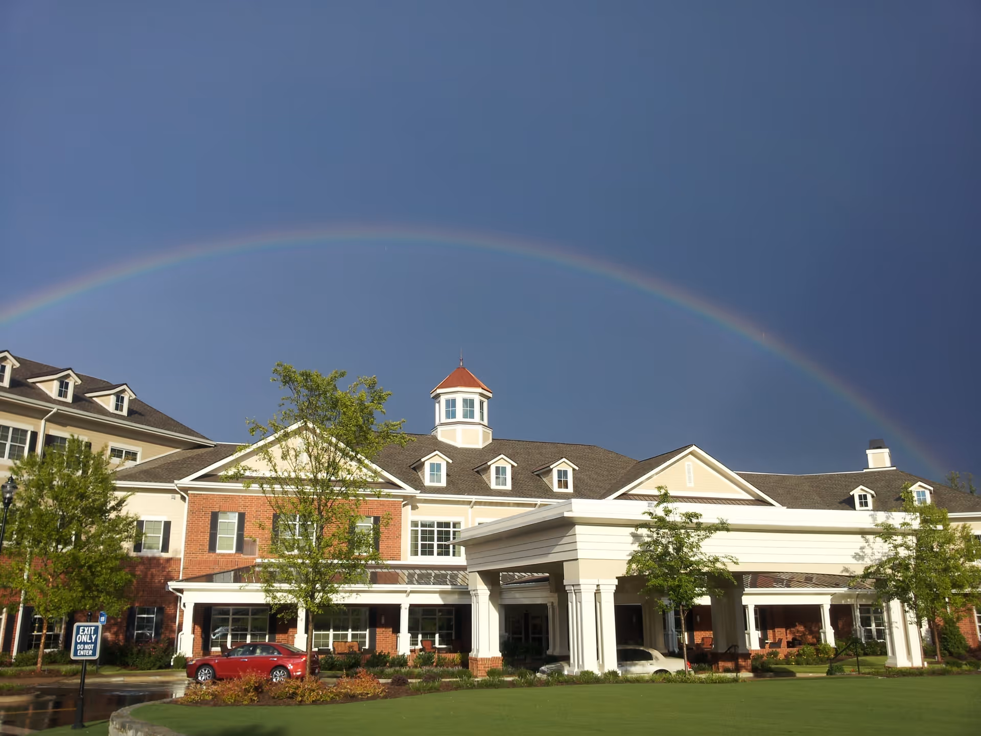 Exterior view of a large brick and beige retirement community building with a covered entrance and multiple windows. A vibrant rainbow arcs across the sky above the building. There are green trees and a well-maintained lawn in front, along with a red car parked near the entrance.