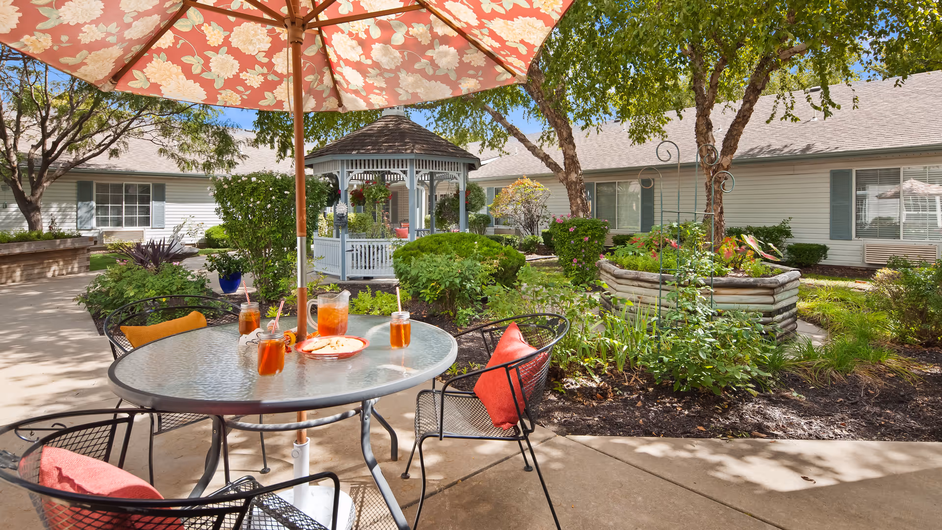Outdoor patio area with a round glass table under a large floral umbrella. Four black metal chairs with red and orange cushions surround the table, which has four glasses of iced tea and a plate with snacks. In the background, there is a white gazebo, green bushes, trees, and a building with white siding and blue shutters.