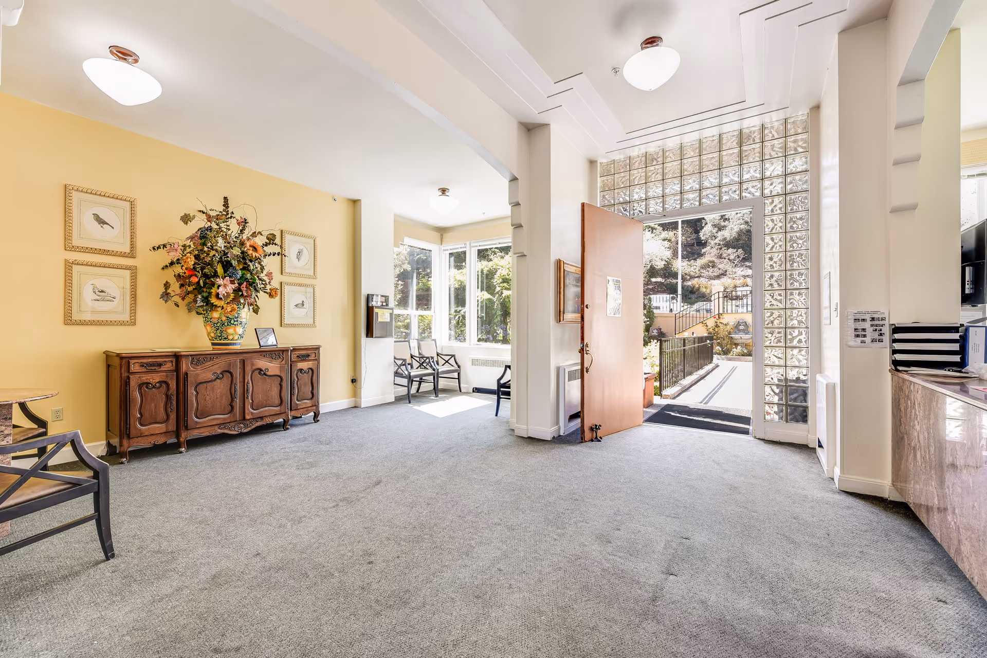 Bright and spacious entryway of Buena Vista Manor House with an open wooden door leading outside. The room features a yellow accent wall with framed bird artwork and a large floral arrangement on a wooden sideboard. There are several chairs near a window letting in natural light, and a reception desk area is visible on the right.