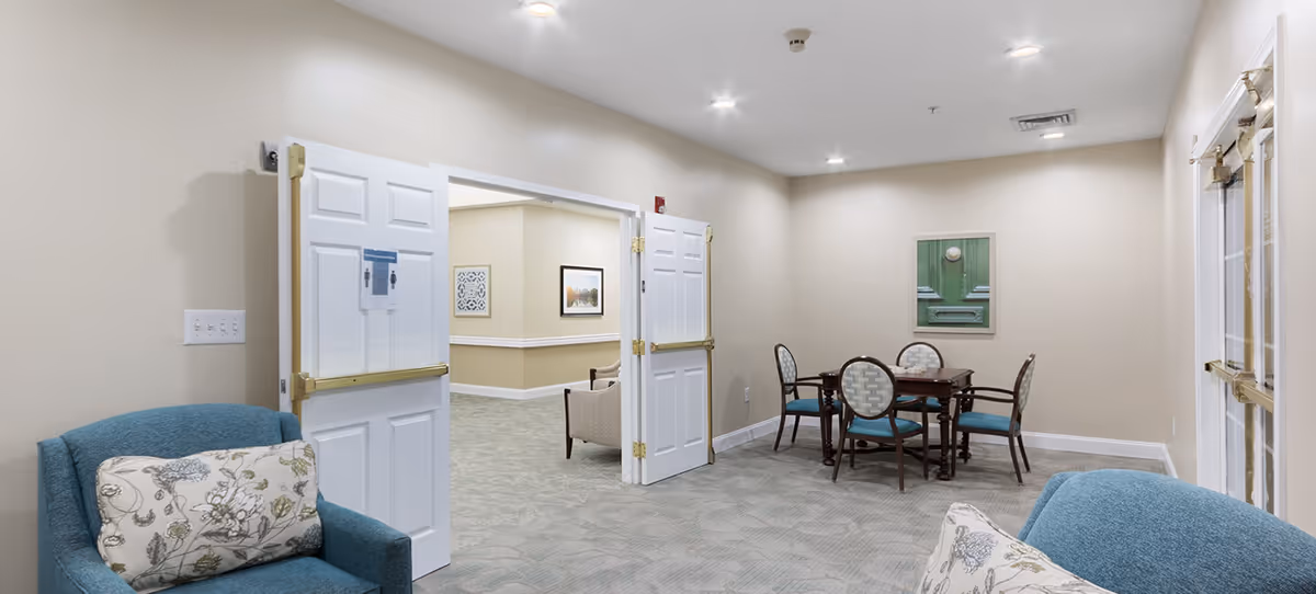 Interior view of a senior living facility lounge area with beige walls and carpeted floor. The room features a small wooden table with four chairs, two blue upholstered armchairs with floral pillows, and double white doors with brass push bars leading to another room with additional seating and framed artwork on the walls.