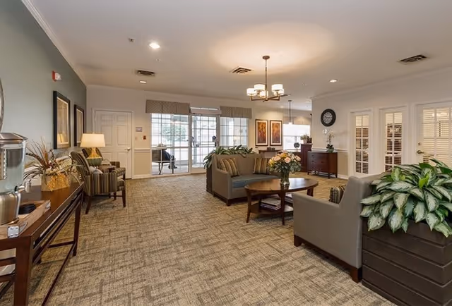 A spacious and well-lit living room area in a senior living facility with comfortable seating including sofas and armchairs, a coffee table with a flower vase, indoor plants, framed artwork on the walls, and large windows allowing natural light to enter.