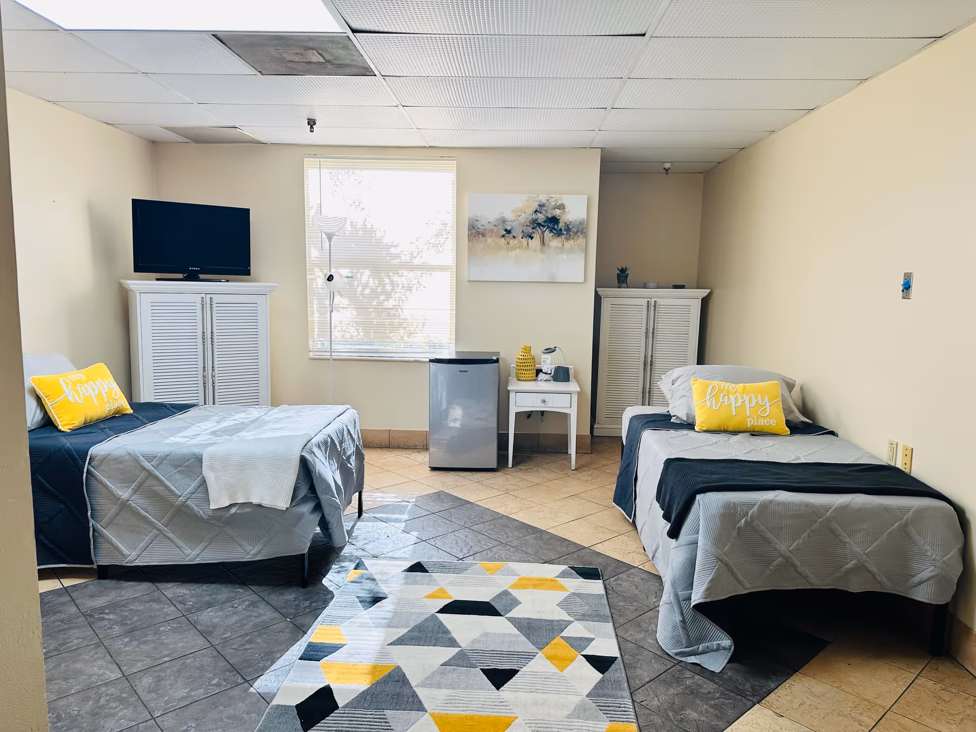 Bright shared bedroom with two twin beds, a TV on a cabinet, a small fridge, nightstand, and decorative pillows.