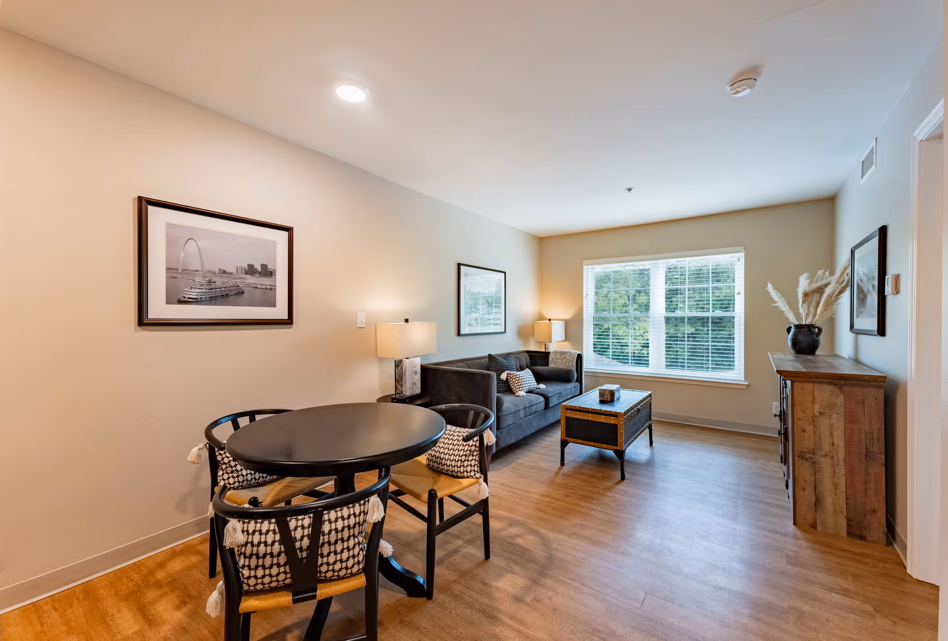 A bright living room in a senior living facility featuring a round black dining table with four chairs, a gray sofa with patterned cushions, a wooden coffee table, two table lamps, framed artwork on the walls, a large window with white blinds, and a wooden sideboard with a decorative vase.