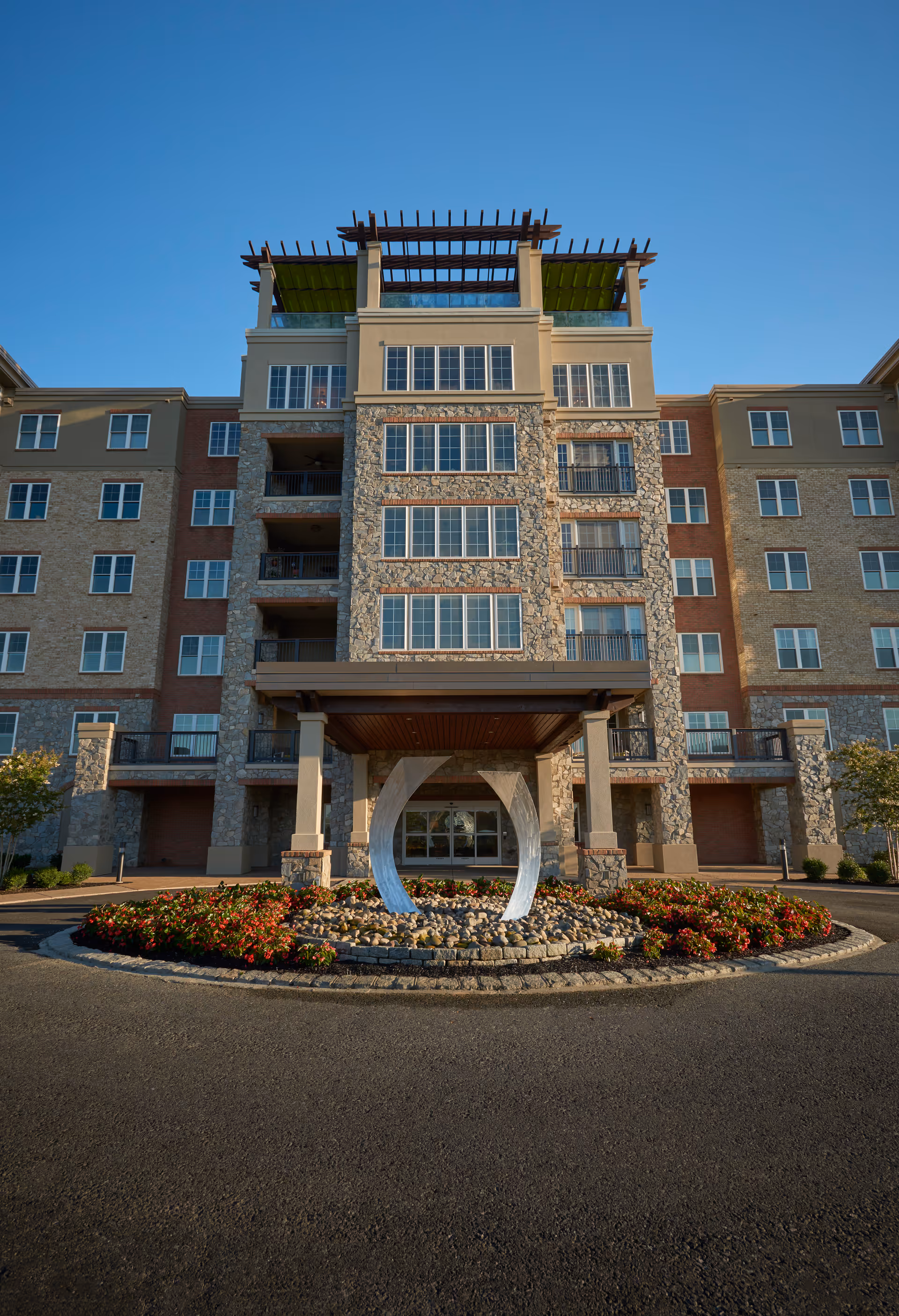 Front exterior view of a multi-story senior living facility building with stone and brick facade, large windows, a covered entrance supported by columns, and a circular flower bed with a modern metal sculpture in front.