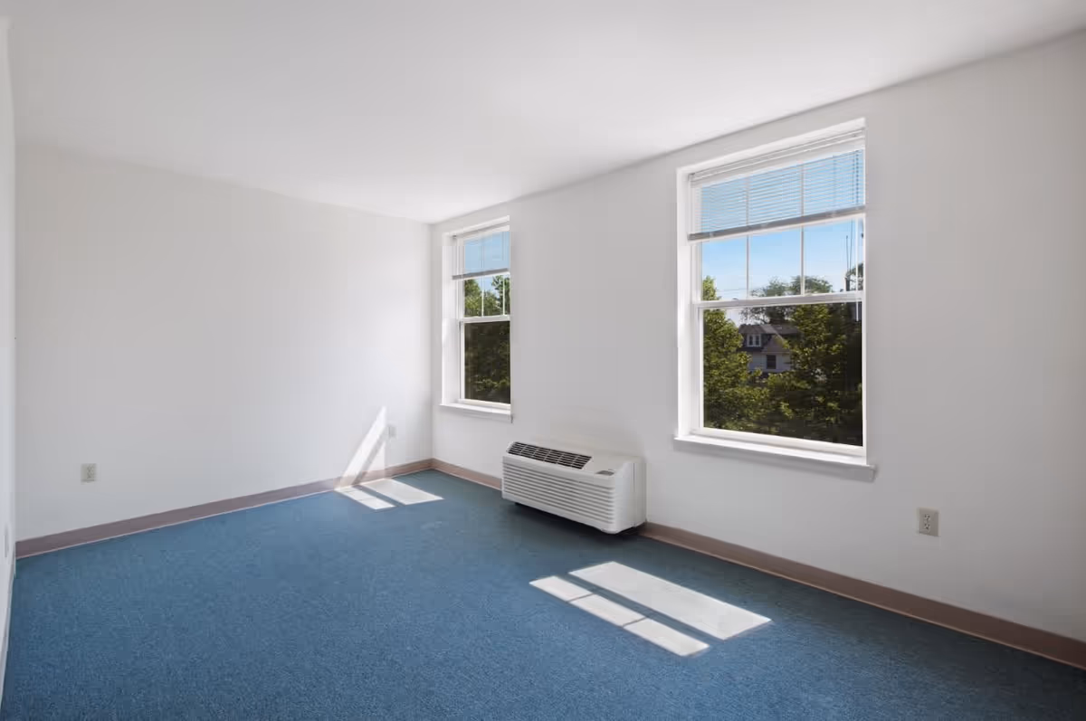 Empty room with white walls, two large windows with blinds partially open, blue carpeted floor, and a white air conditioning unit beneath one window. Sunlight casts shadows on the floor.
