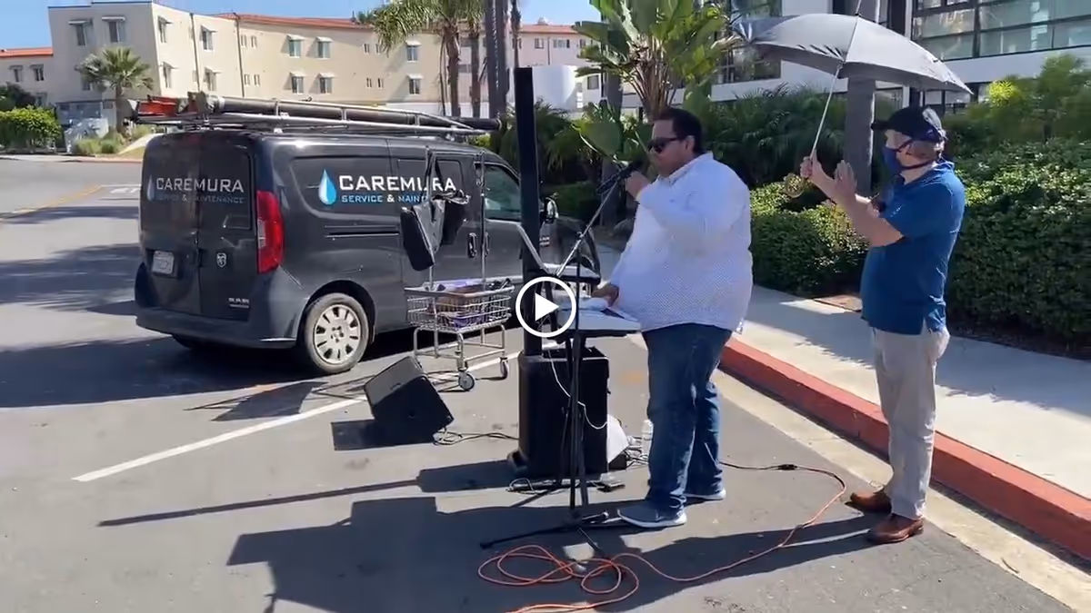 Two men standing outside near a black Caremura service van. One man is speaking into a microphone at a stand, while the other man holds an umbrella over him. They are on a paved area with buildings and greenery in the background.