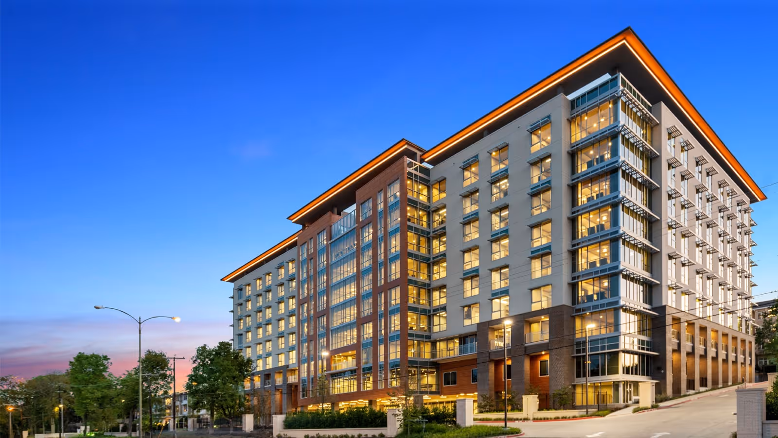 Wide-angle view of a modern multi-story residential building illuminated at dusk with numerous windows and landscaped grounds.