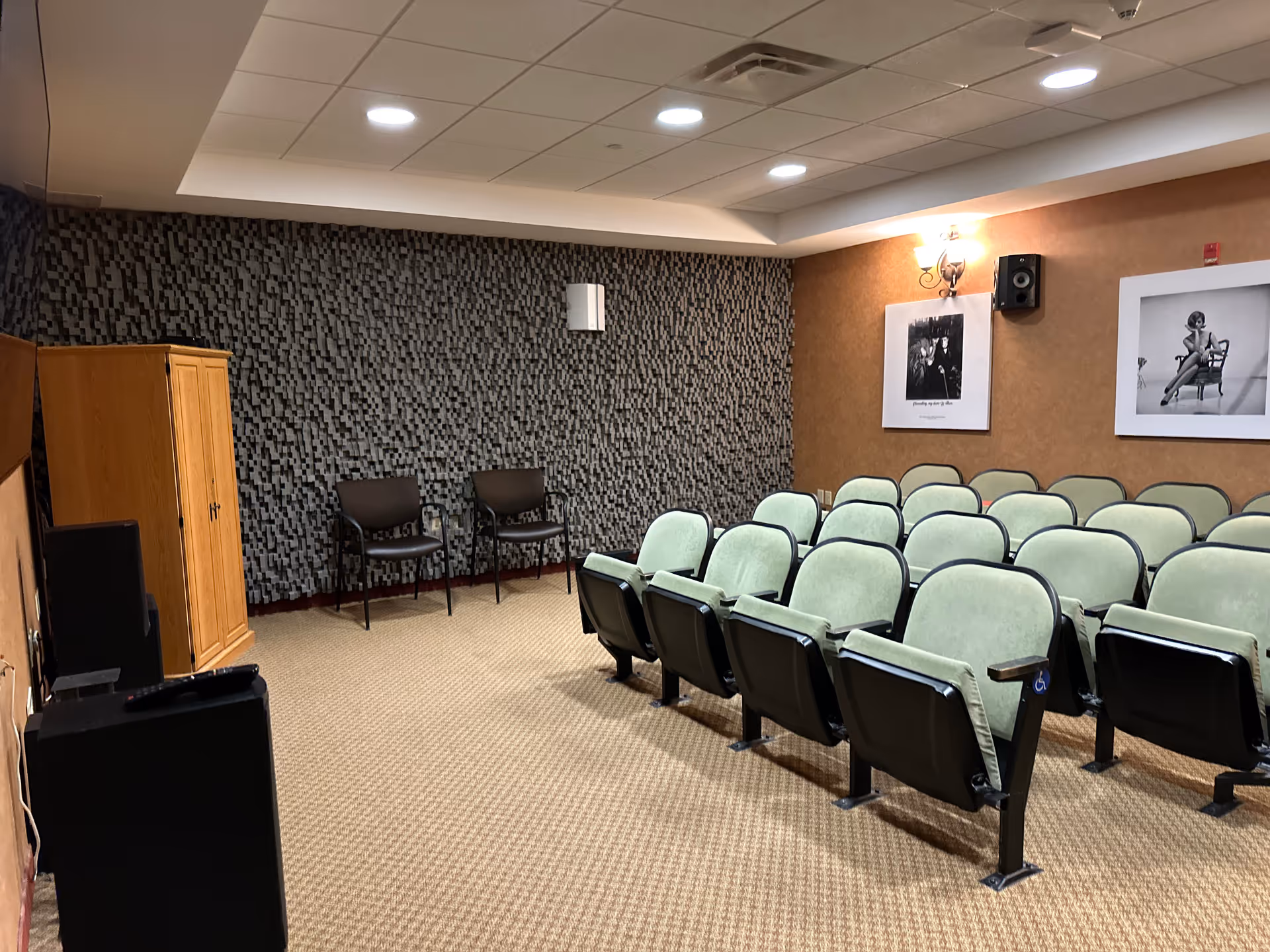 A small theater or presentation room with rows of green cushioned seats facing a wall with two black and white framed photographs. The room has a patterned carpet, a textured wall, two black chairs in the corner, a wooden cabinet, and ceiling lights.