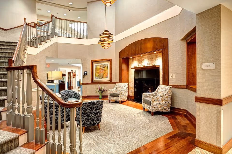 A cozy interior common area with a staircase on the left, two patterned armchairs facing a fireplace with a wooden mantel, a beige carpet, and a hanging light fixture. There is a framed abstract painting on the wall and a vase with flowers on a small table.