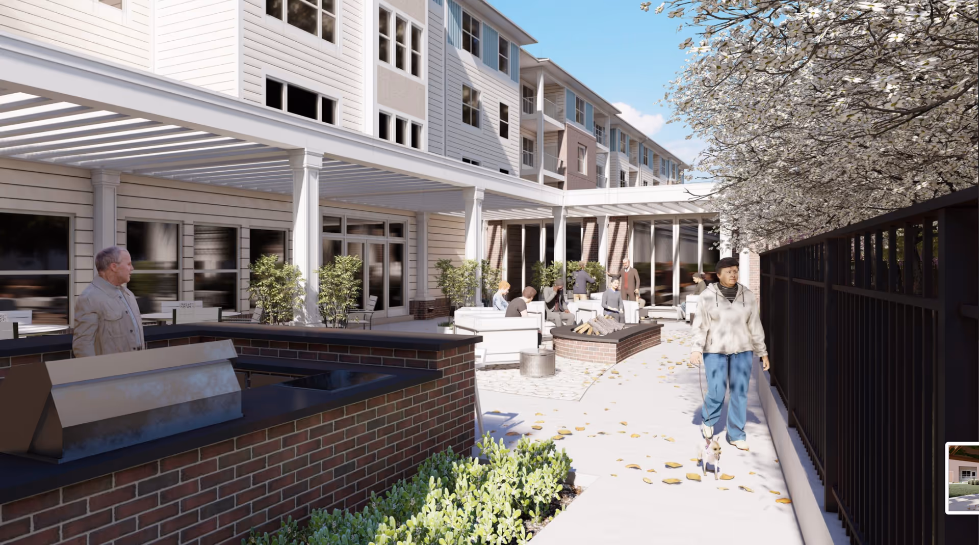 Outdoor patio area at Acclaim at Cary Pointe featuring a brick barbecue grill, white cushioned seating around a fire pit, several people sitting and standing, and a person walking a small dog along a pathway lined with flowering trees and a black metal fence.