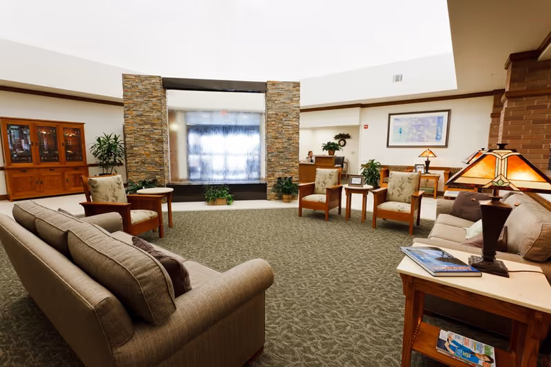A spacious senior living facility lounge area with comfortable sofas and armchairs arranged around a central carpeted space. There is a decorative stone water feature in the background, wooden furniture including side tables and a cabinet, potted plants, and warm lighting from table lamps. The room has a welcoming and cozy atmosphere.