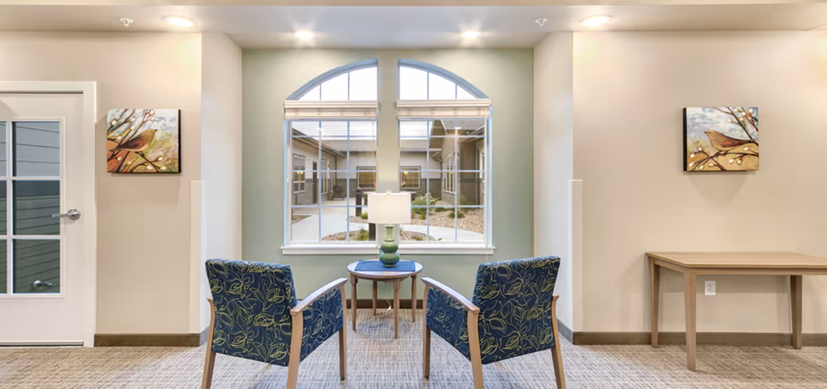 Two upholstered chairs and a small table with a lamp arranged in front of a large arched window overlooking an interior courtyard.