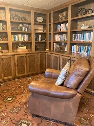 A cozy reading nook featuring a brown leather armchair with a decorative pillow, positioned in front of wooden built-in bookshelves filled with books and decorative items. The floor is covered with a patterned carpet.