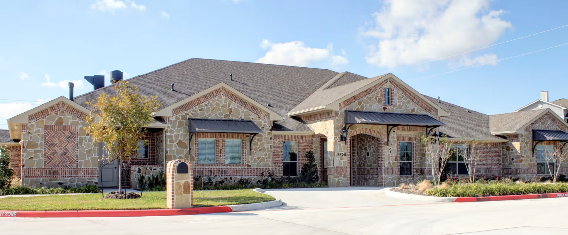 Exterior view of a single-story building with stone and brick facade, multiple windows with black awnings, a small tree and landscaping in front, and a driveway with a red curb labeled 'FIRE LANE'.