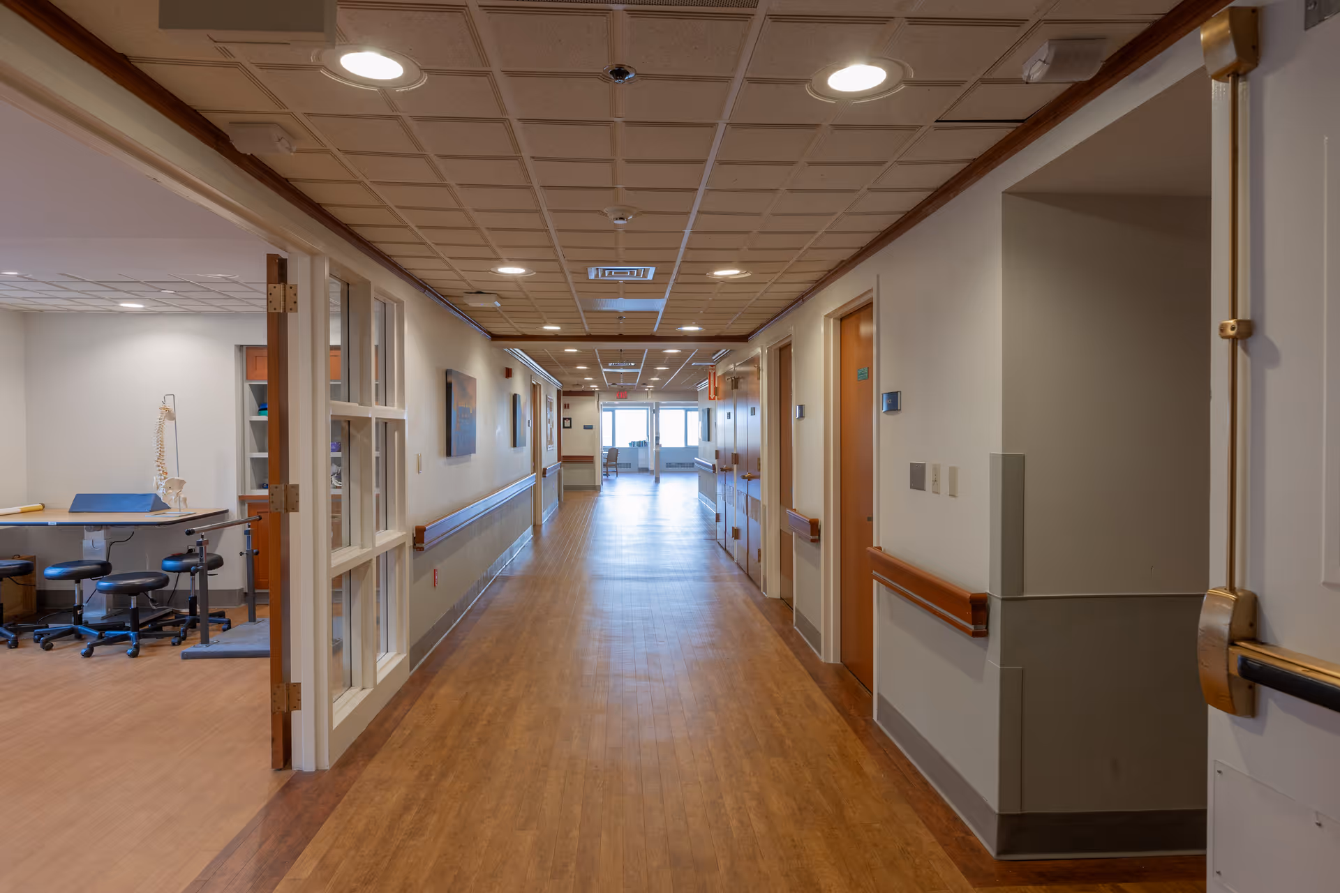 A long, well-lit hallway in a healthcare facility with wooden floors and handrails on both sides. On the left, there is an open room with medical stools, a table, and a model of a human spine. Several closed doors line the right side of the hallway, and there is natural light coming from windows at the far end.