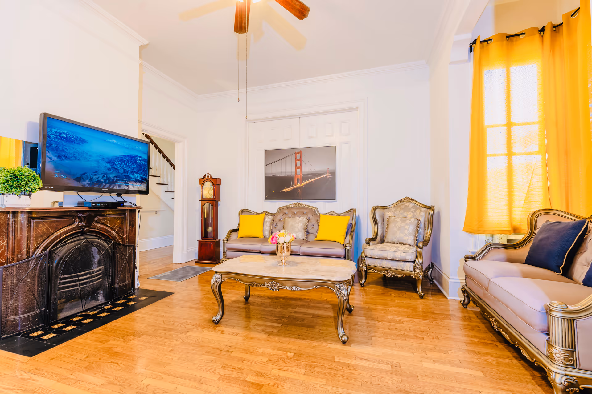 A bright living room with wooden flooring, featuring a vintage-style sofa set with yellow and dark blue cushions, a marble-top coffee table, a wall-mounted TV above a dark fireplace, a grandfather clock, and yellow curtains on the window.
