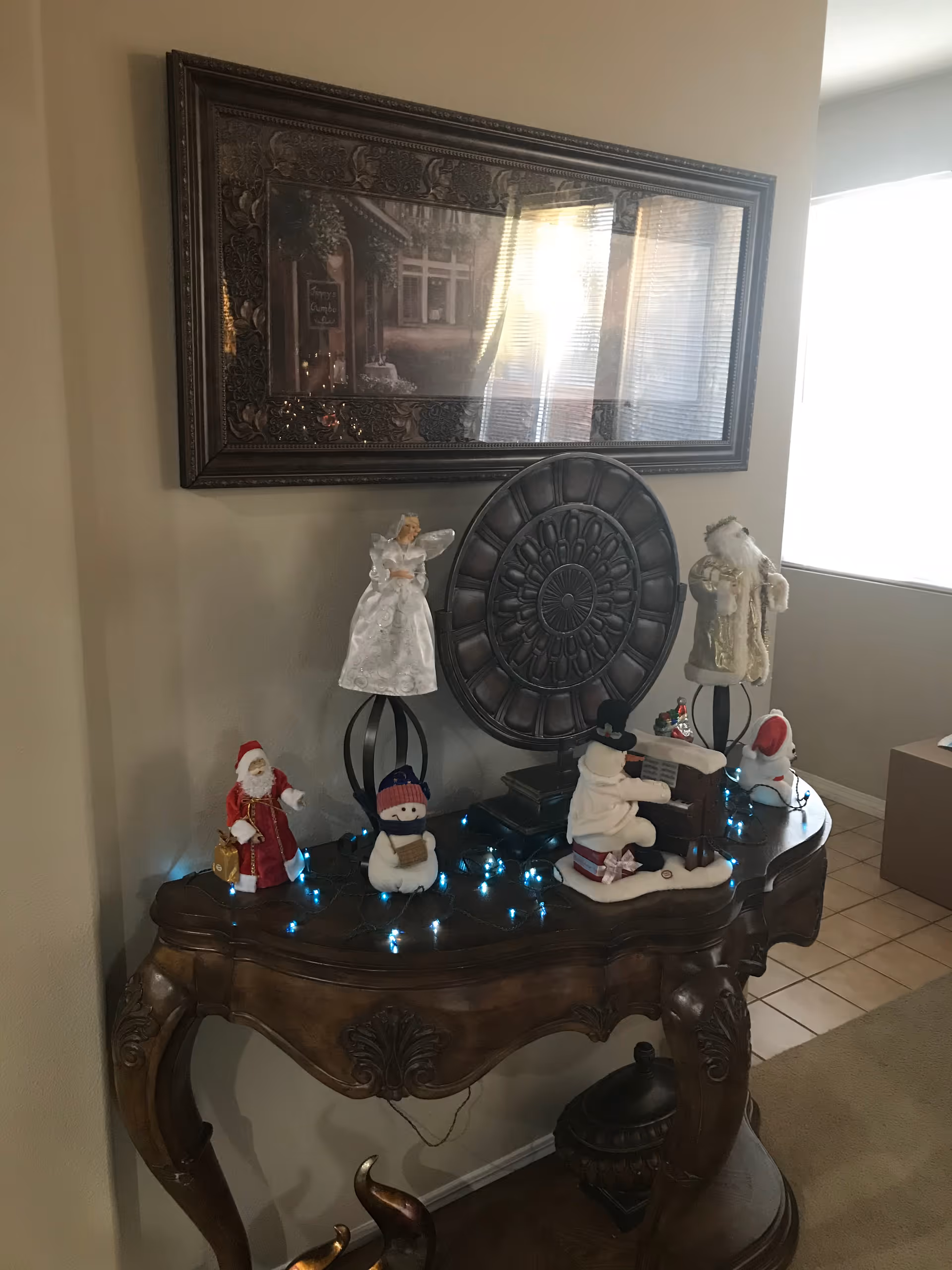 A decorative wooden console table with curved legs and ornate carvings, displaying various Christmas-themed figurines including a snowman, Santa Claus, an angel, and a snowman playing a piano. Blue string lights are draped across the table. Above the table is a large framed artwork with a reflective glass surface. The setting appears to be an interior corner of a room with tiled flooring and a window letting in natural light.