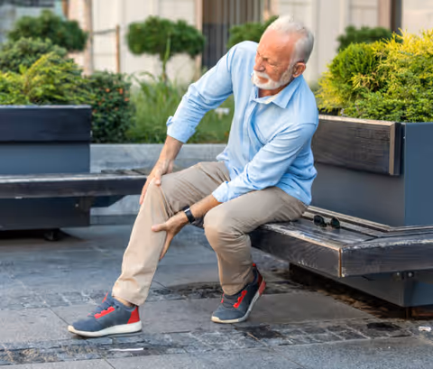 An elderly man with white hair and beard sitting on a wooden bench outdoors, holding his lower leg and appearing to be in pain. He is wearing a light blue shirt, beige pants, and gray sneakers with red accents. There are green bushes and plants in the background.