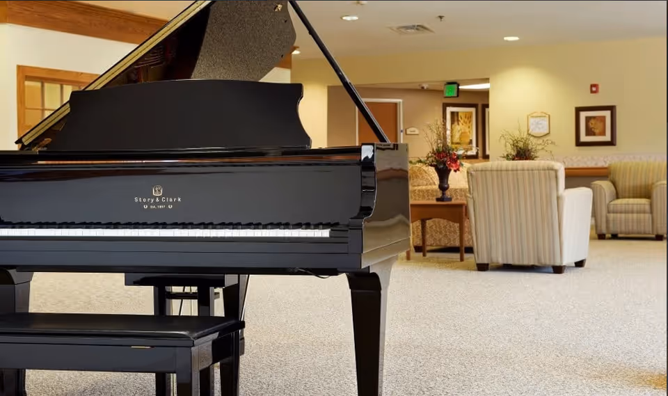 A black Story & Clark grand piano with a matching bench in a spacious room featuring beige carpet, several upholstered armchairs, a wooden coffee table with a floral arrangement, and framed artwork on the walls.