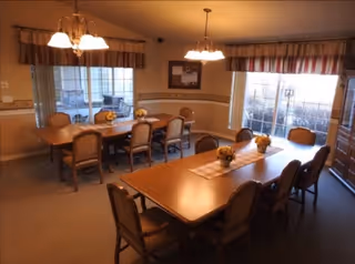 Interior dining room with two long wooden tables and chairs, hanging light fixtures, and windows with striped valances.