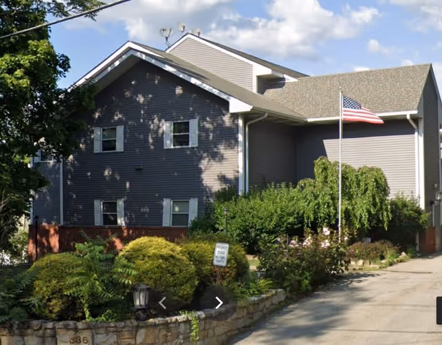 Exterior view of a two-story residential building with gray siding and a gable roof. The building is surrounded by green shrubs and trees, with an American flag on a flagpole near the entrance. A stone retaining wall lines the driveway leading to the building.