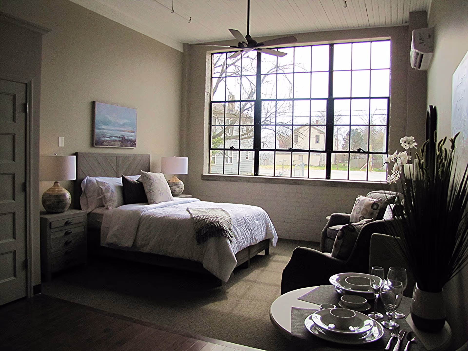 Bright bedroom with a bed, nightstands, armchair and a small dining table in front of a large multi-pane window.