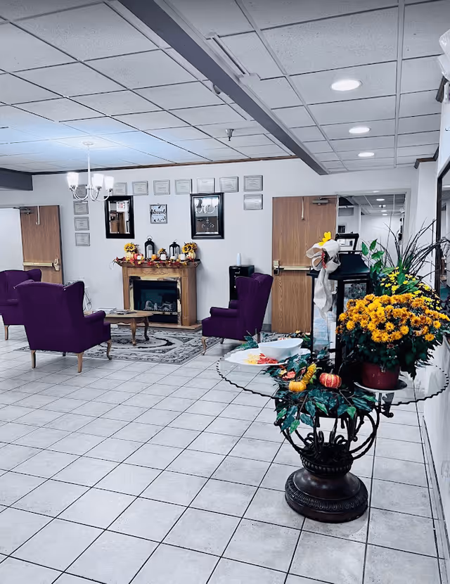 A cozy common area in an assisted living facility featuring a tiled floor, a glass table with a floral arrangement and decorative items, purple upholstered armchairs arranged around a wooden coffee table, a fireplace with fall-themed decorations, framed certificates and mirrors on the wall, and wooden doors in the background.