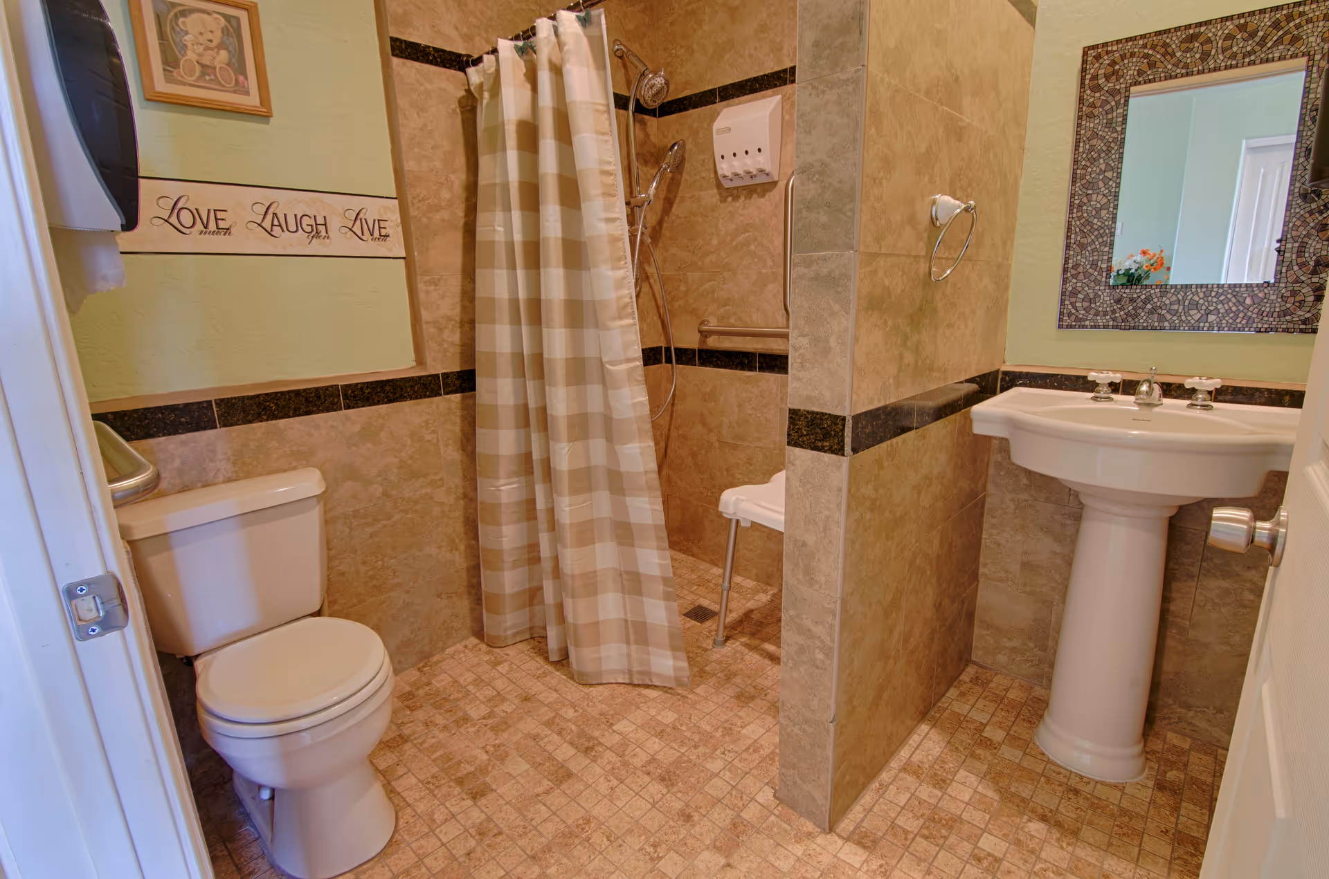 A bathroom with beige tiled walls and floor, featuring a white toilet, a pedestal sink with a decorative framed mirror above it, and a walk-in shower with a beige and white checkered shower curtain and a white shower chair inside. There is a wall decoration with the words 'Love Laugh Live' and a framed picture of a teddy bear on the wall.