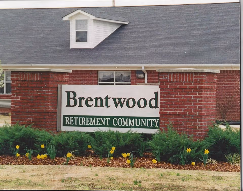 A brick sign structure with a white sign that reads 'Brentwood Retirement Community' in front of a building with a gray roof and a small dormer window. There are green bushes and yellow flowers planted in front of the sign.