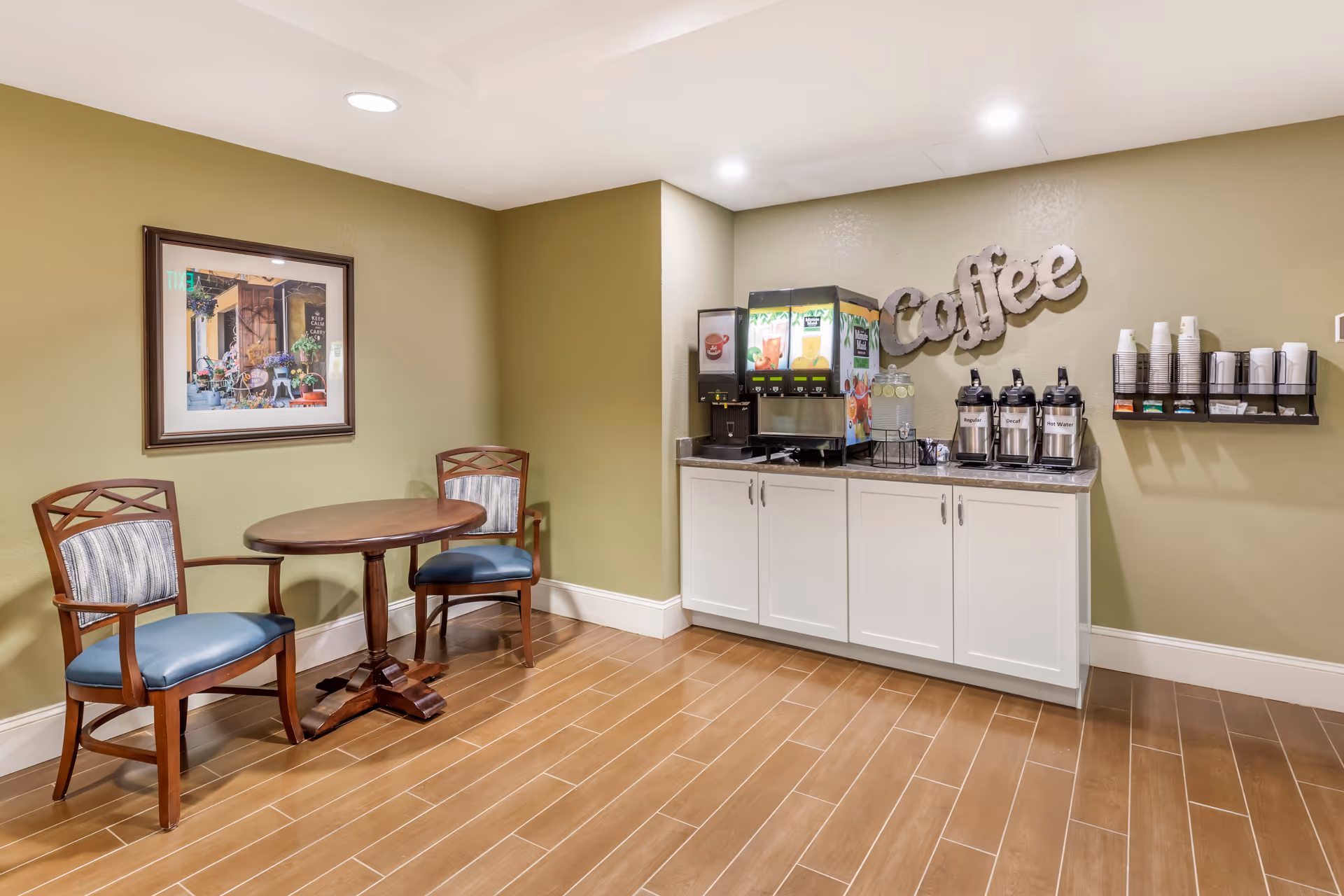 A small coffee and beverage station area with a wooden table and two chairs with blue cushions. The station includes coffee dispensers, juice machines, cups, and a sign on the wall that says 'Coffee'. The walls are painted light green and the floor has brown tiles.