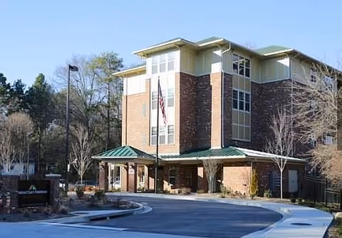 Exterior view of a multi-story senior living facility building with a brick and beige facade, green roof accents, and an American flag displayed near the entrance. The building is surrounded by trees and a curved driveway leading to the entrance.
