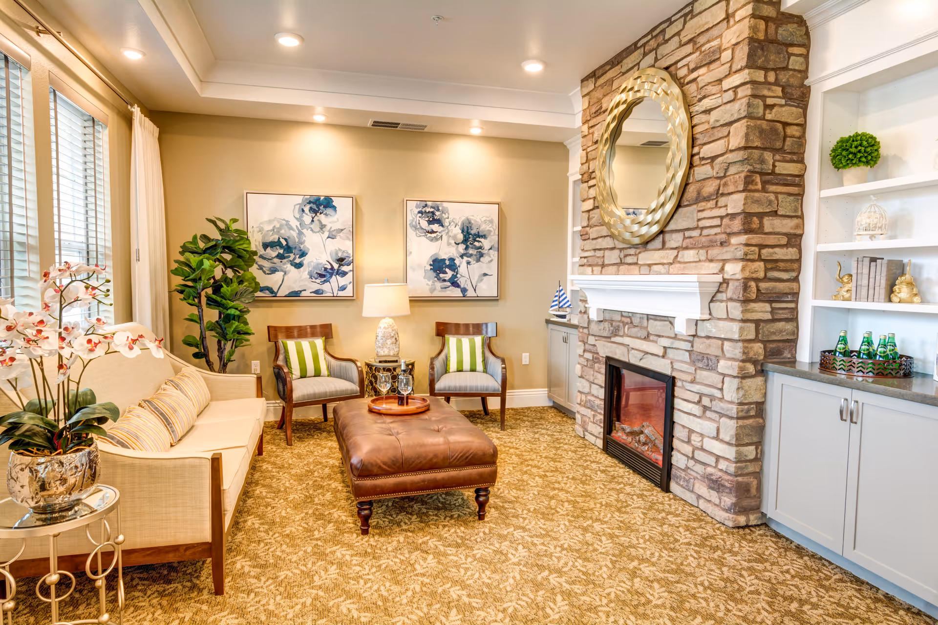 A cozy living room with a beige sofa, two wooden chairs with green and white striped cushions, a brown leather ottoman, and a stone fireplace with a round decorative mirror above it. The room features floral artwork on the wall, a potted plant, built-in shelves with decorative items, and a window with white curtains allowing natural light in.