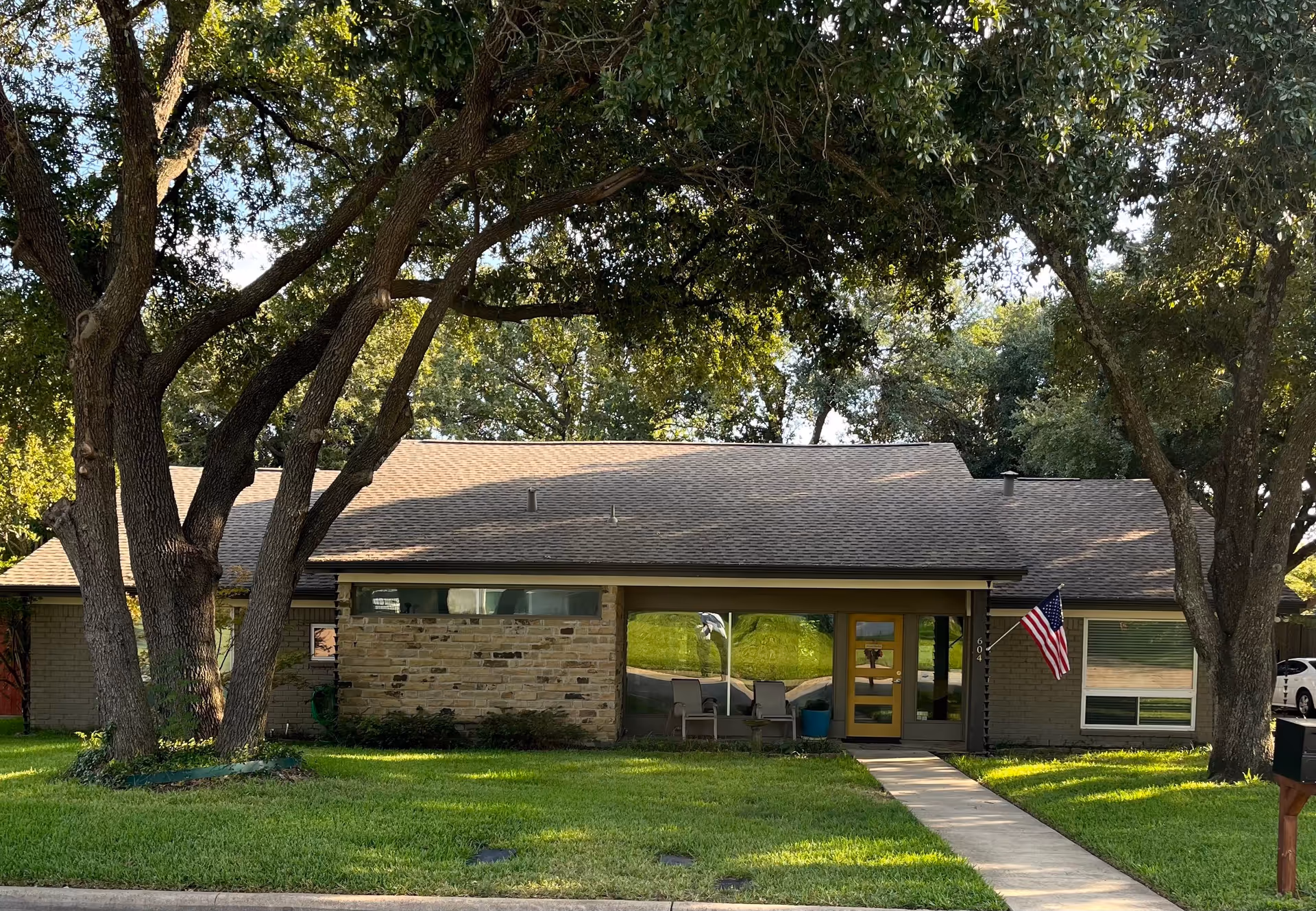 Single-story residential building with a sloped roof, large windows, and a yellow front door. The building is surrounded by green grass and large trees providing shade. An American flag is mounted near the entrance, and two chairs with a small table are placed on the porch.