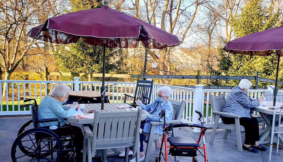 Three elderly women wearing masks sitting at outdoor tables under purple umbrellas, engaged in activities such as writing or drawing. One woman is in a wheelchair, and a red walker is nearby. The setting is a fenced patio area with trees and greenery in the background.