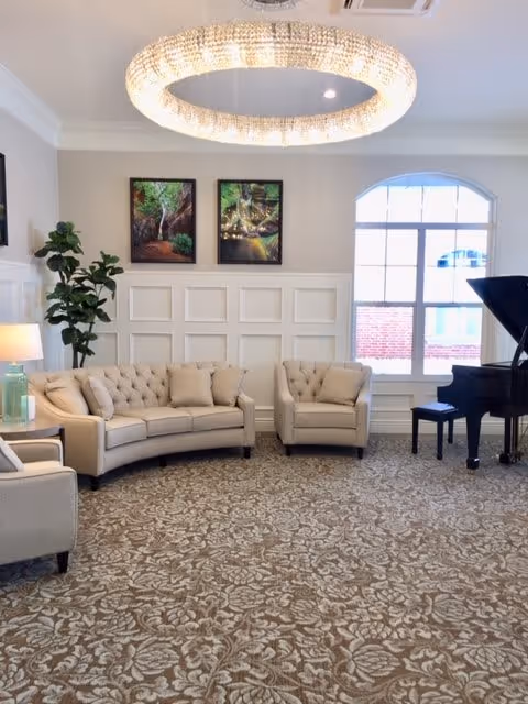 A bright and elegant living room with a large window letting in natural light. The room features a beige curved sofa, a matching armchair, a grand piano with a bench, a tall green plant, two framed nature-themed paintings on the wall, a table lamp with a green base, and a large circular chandelier hanging from the ceiling. The floor is covered with a patterned carpet.