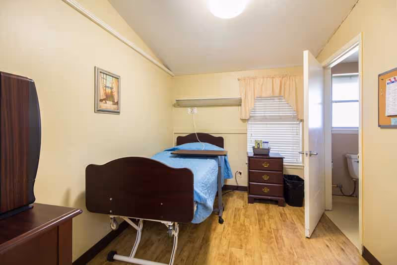 A small, neatly organized bedroom in a senior living facility with a bed, dresser, and open bathroom door.