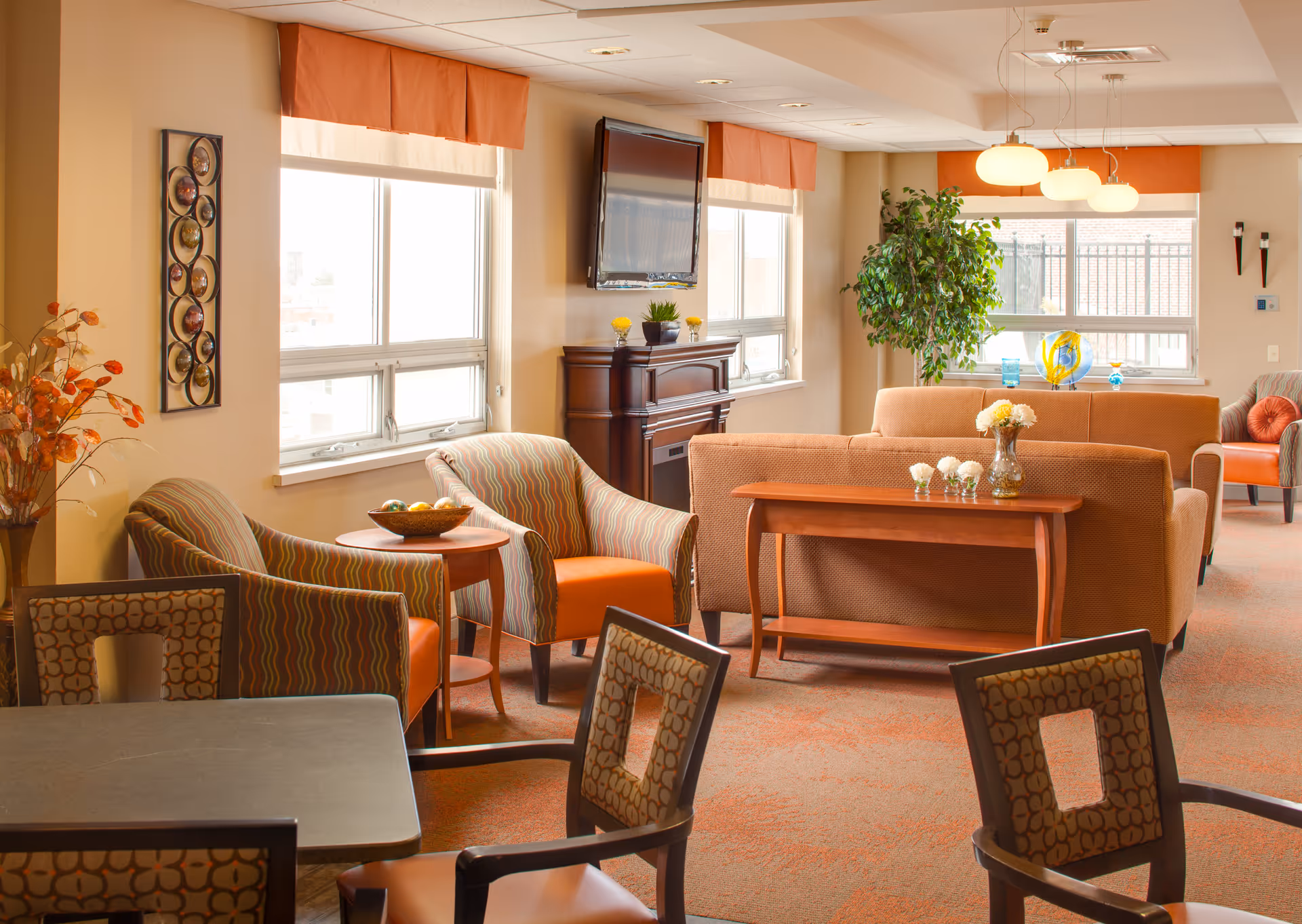 A bright and cozy living room area in a senior living facility with multiple comfortable chairs and sofas in warm tones of orange and brown. There are large windows with orange valances letting in natural light, a wall-mounted TV above a wooden cabinet, a small table with a bowl of fruit, and decorative plants and flowers placed around the room.