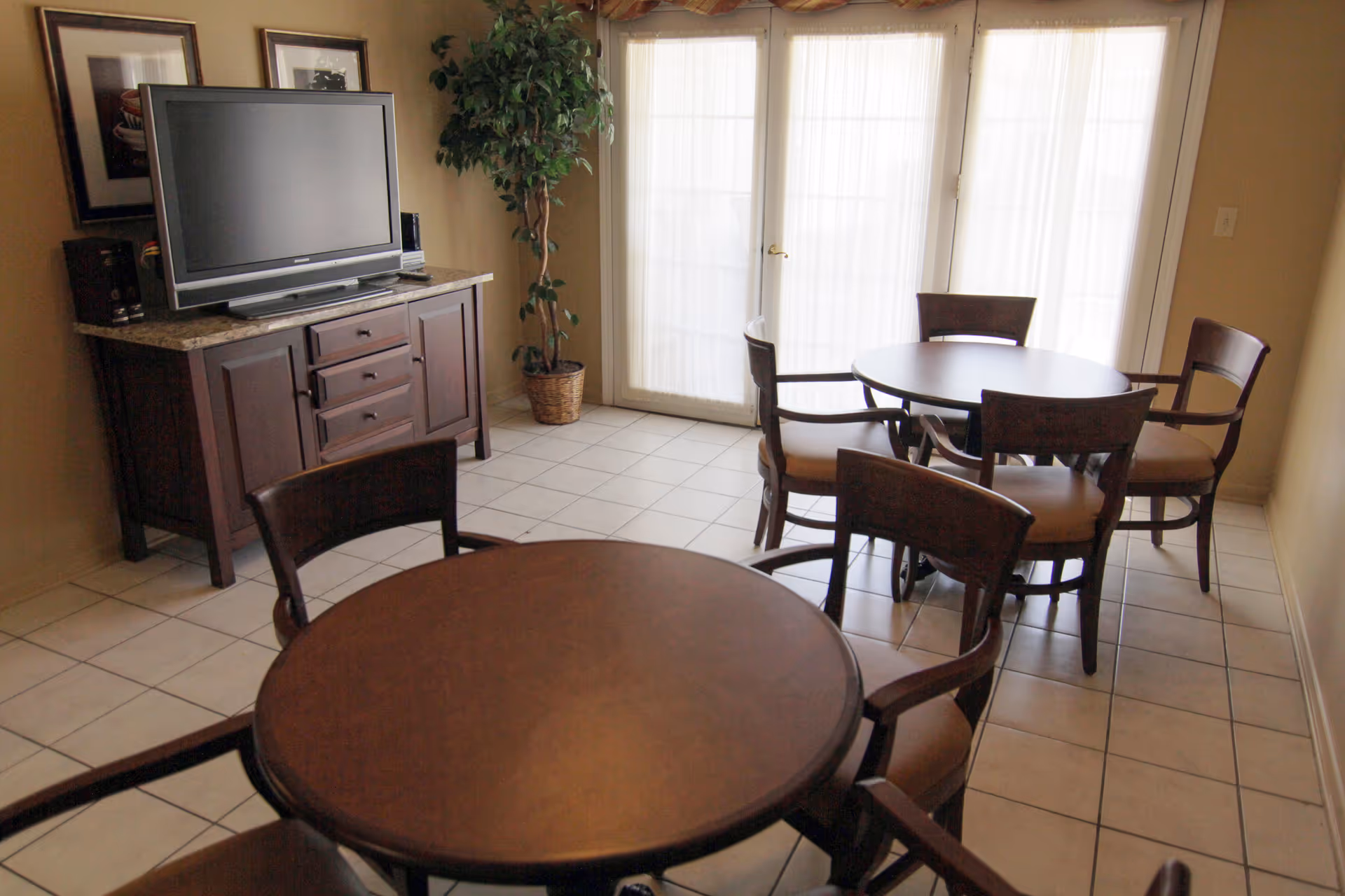 Interior room with two round wooden tables each surrounded by four wooden chairs with cushioned seats. A flat-screen TV sits on a wooden cabinet with drawers and doors. There is a potted plant in the corner near large glass doors covered with sheer white curtains.