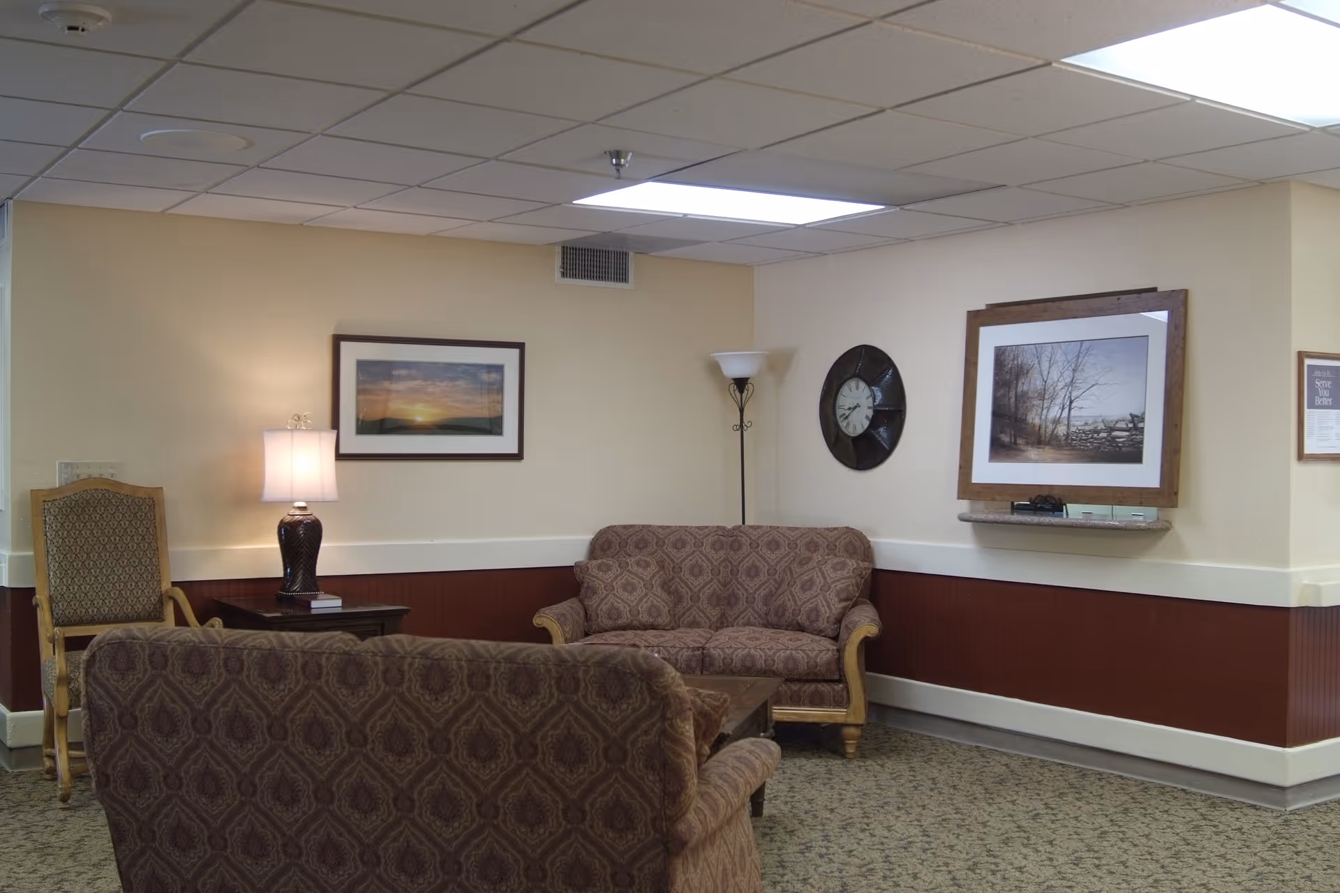 A nursing-home common room with upholstered sofas and chairs, framed pictures, lamps, and a wall clock.
