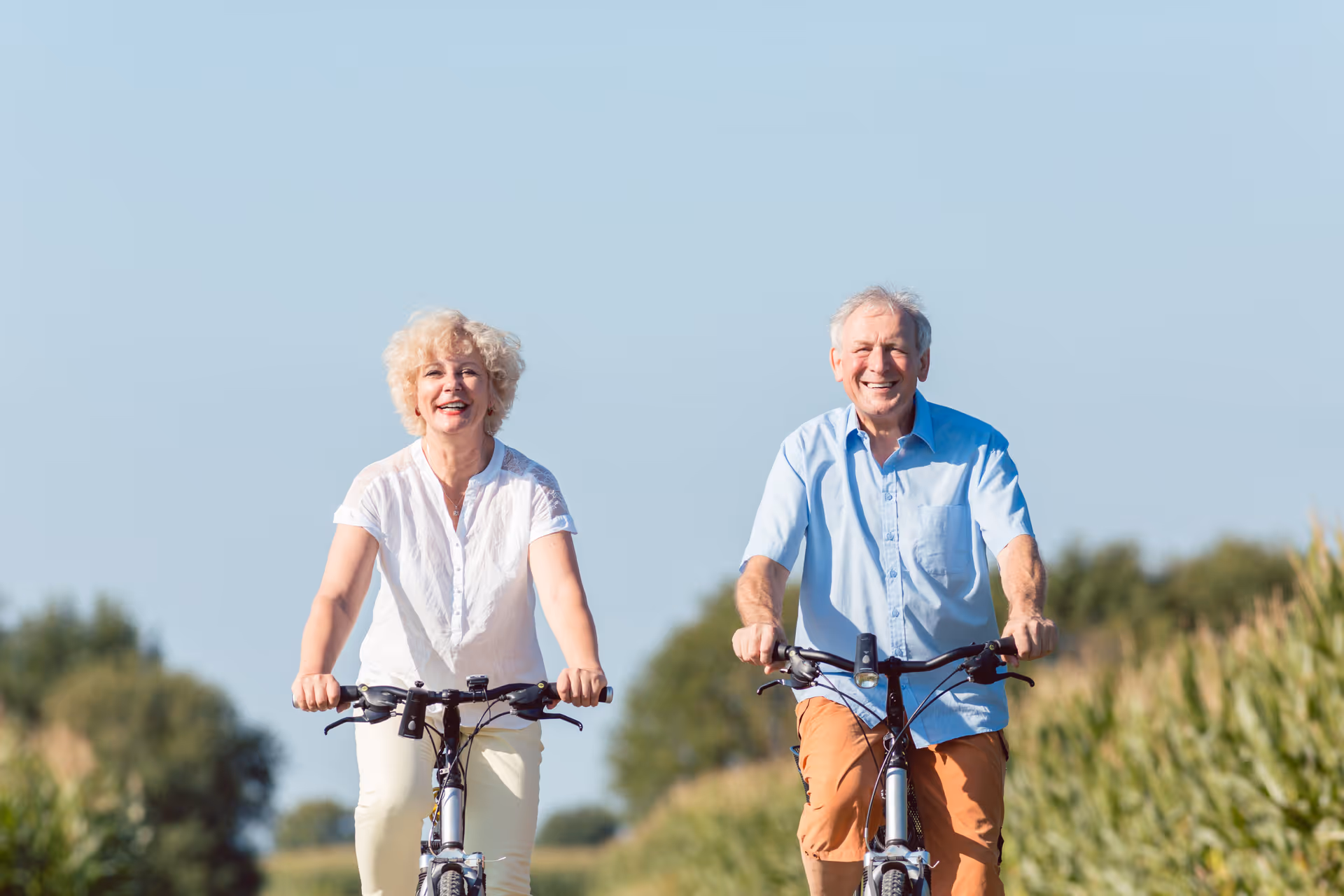 An elderly woman and man riding bicycles outdoors on a sunny day with greenery and blue sky in the background, both smiling and enjoying the ride.