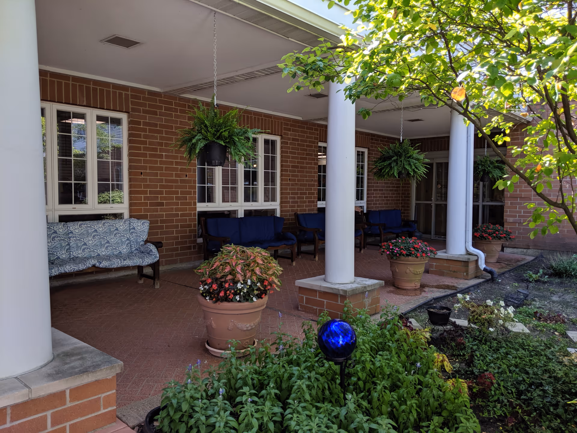 Covered outdoor patio area with brick walls and white columns, featuring hanging green plants, potted flowers, and cushioned benches. There is a garden with green plants and a blue decorative orb in the foreground.