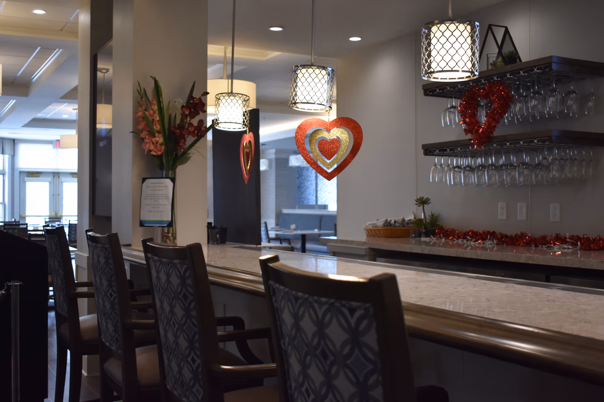 Interior view of a bar area in a senior living facility with a marble countertop, several high-backed chairs, hanging pendant lights, and heart-shaped decorations. Shelves with hanging wine glasses and a vase with flowers are visible, along with a dining area in the background.