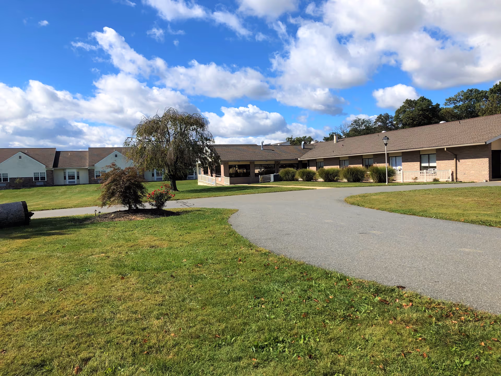 Exterior view of a single-story retirement community building with a paved driveway, green lawn, and a few trees under a partly cloudy blue sky.