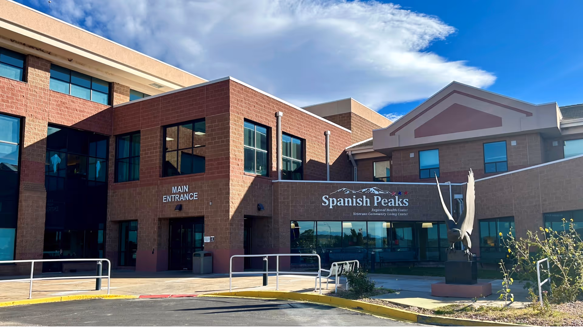 Exterior view of the Spanish Peaks Veterans Community Living Center building showing the main entrance with large windows and a sculpture of an eagle with wings spread in front. The building is made of brick and has multiple stories under a partly cloudy sky.